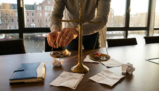 Handen die een messing weegschaal balanceren met symbolische objecten op een vergadertafel, omringd door onderzoeksnotities in warm kantoorlicht.