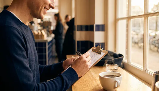 Glimlachende klant vult feedbackformulier in op klembord in Amsterdams café, dampende koffie en warm middaglicht op de achtergrond.